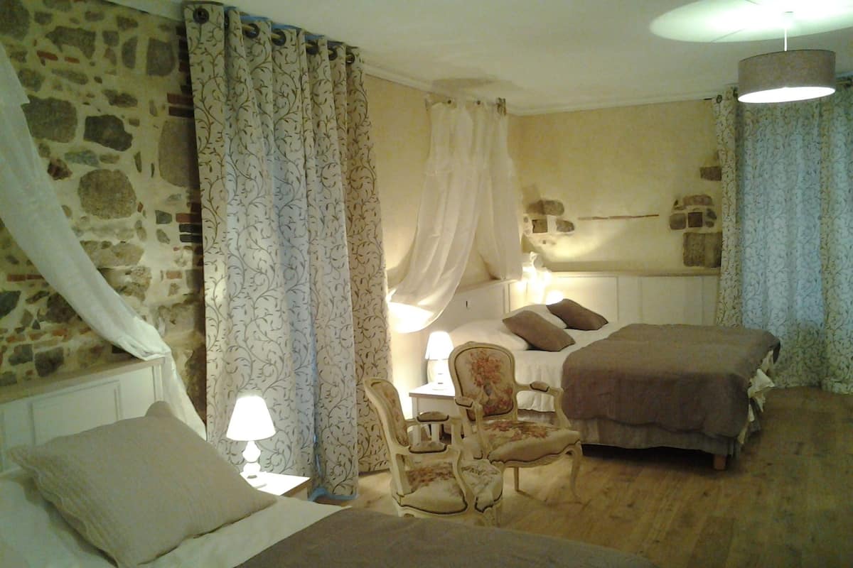 A bedroom with a stone wall, a bed with a canopy, a bedside table with a lamp, a chair, and a second bed.
