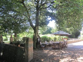 Un área de comedor al aire libre con una mesa y sillas bajo un gran paraguas, rodeada de árboles y un muro de piedra.
