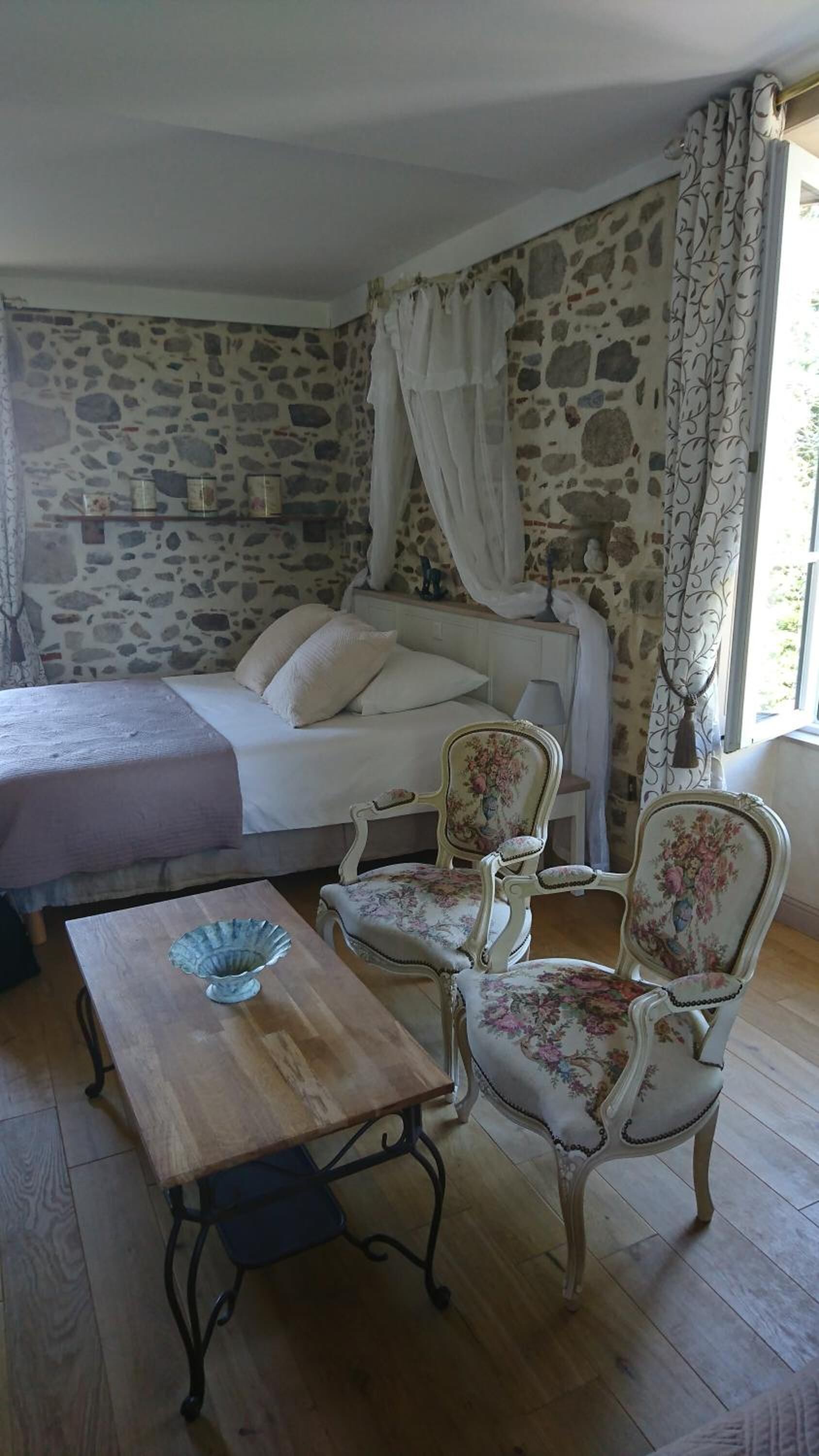 A bedroom with a stone wall, a bed with white bedding, a wooden table with a blue bowl, and two upholstered chairs with floral upholstery.