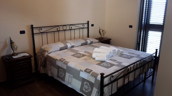 Una cama bien hecha con una colcha estampada y toallas enrolladas, una mesita de noche con lámpara y una ventana con persianas.