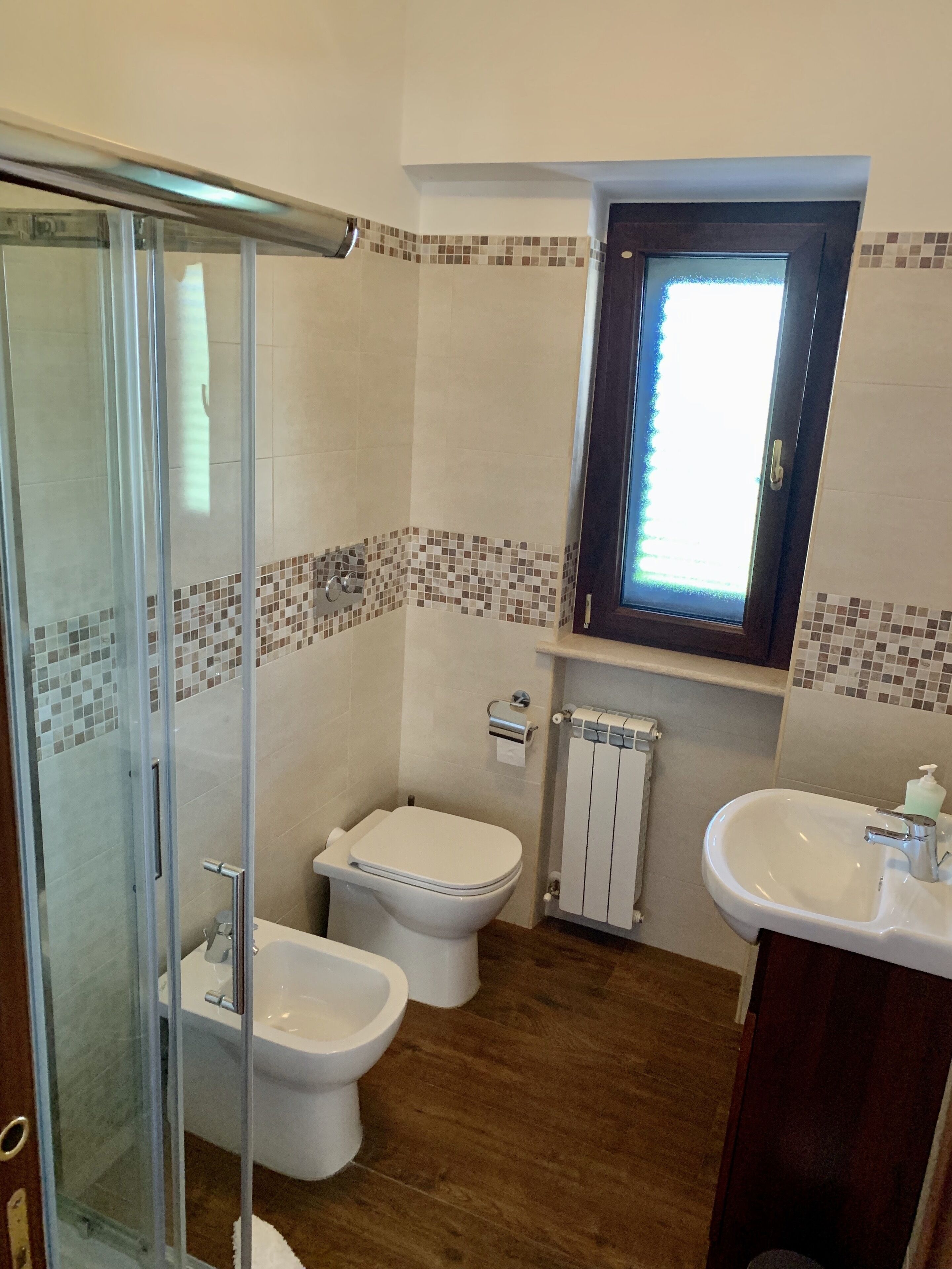 A bathroom with a glass shower enclosure, two toilets, a sink, and a window.