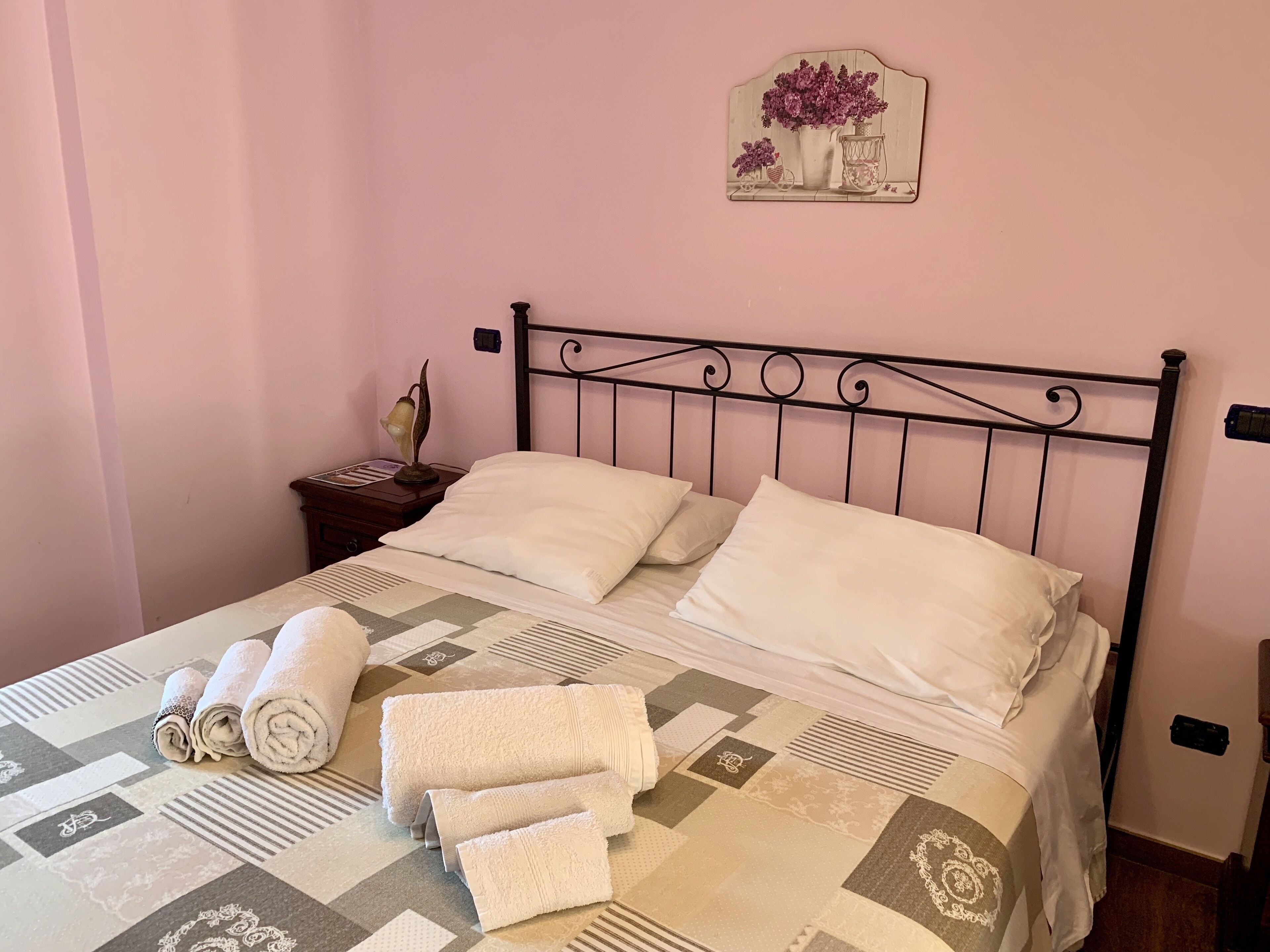A neatly made bed with white linens and rolled towels, a decorative wall piece above the bed, and a nightstand with a lamp and a small plant.