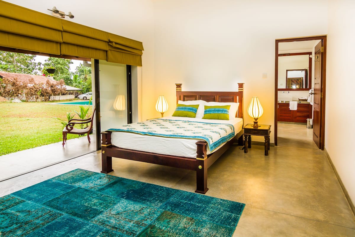 A bedroom with a bed, bedside lamps, a view of the garden, and a bathroom visible through an open door.