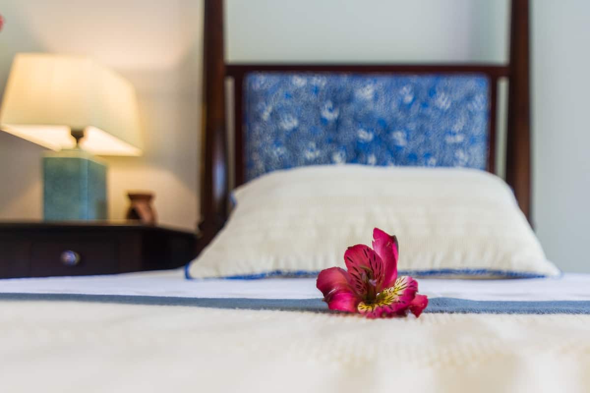 A bed with a white pillow and a single pink flower on the bedspread.
