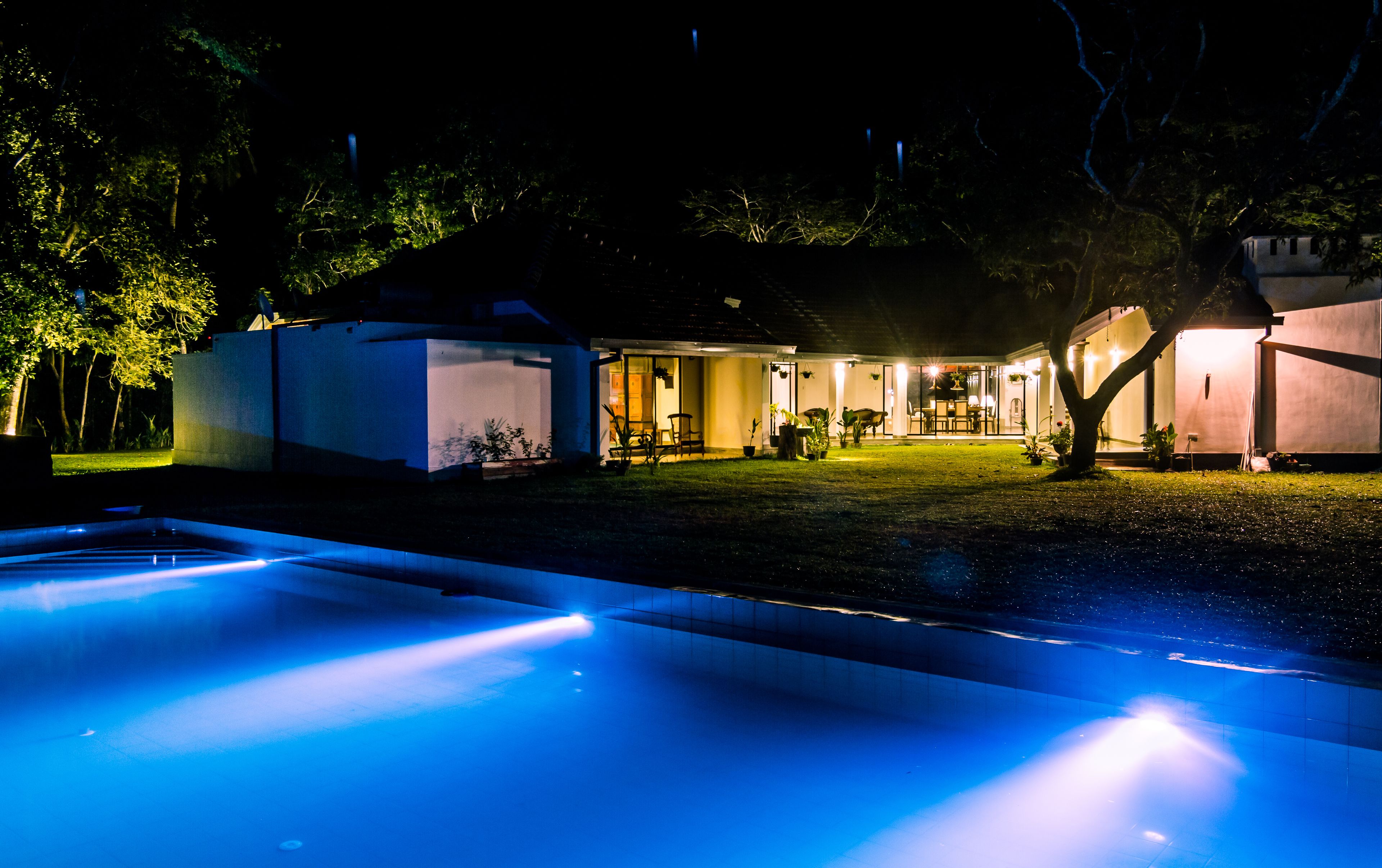 A nighttime view of a residential property with a swimming pool, a covered patio, and a well-lit backyard.