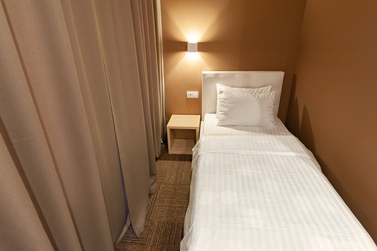 A hotel room with a single bed, a nightstand, a wall-mounted lamp, and a curtain.