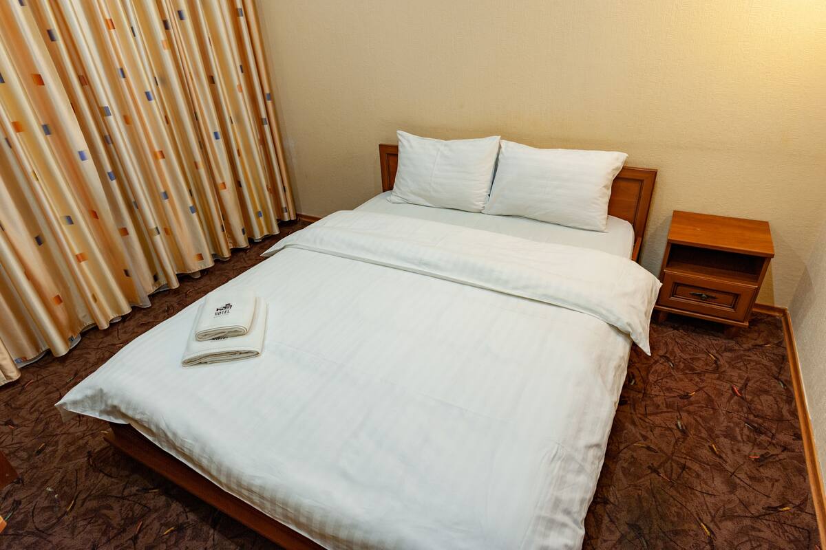 A neatly made bed with white linens, a wooden headboard, and a small wooden nightstand beside it.