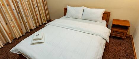 A neatly made bed with white linens, a wooden headboard, and a small wooden nightstand beside it.
