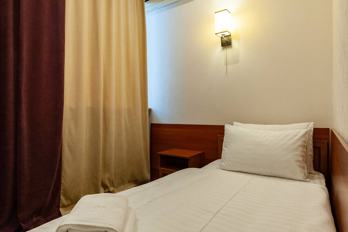 A hotel room with a bed, a nightstand, a wall-mounted light, and a window with curtains.