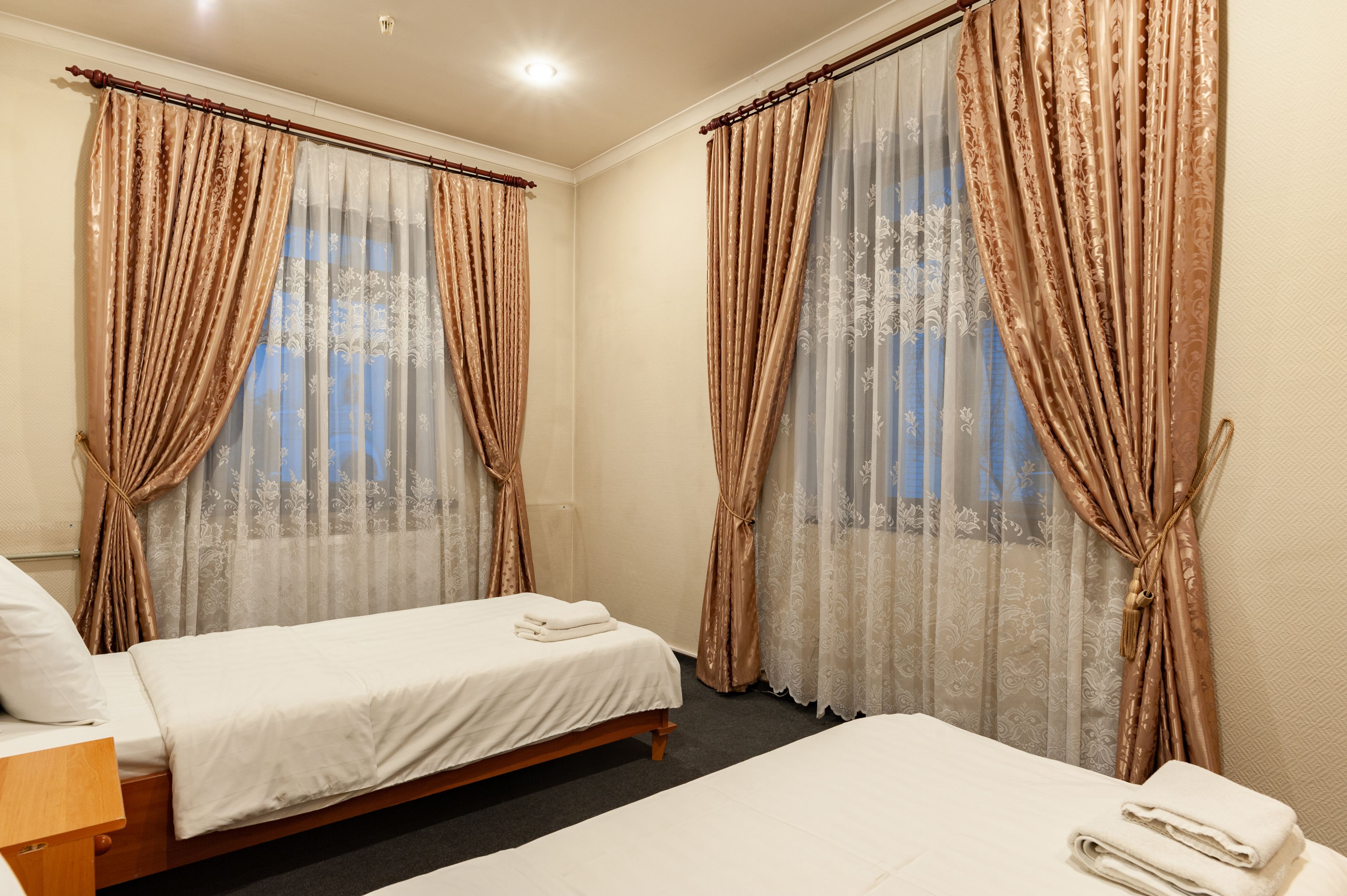 A hotel room with two single beds, a wooden headboard, a small bedside table, and a window with sheer curtains and patterned drapes.