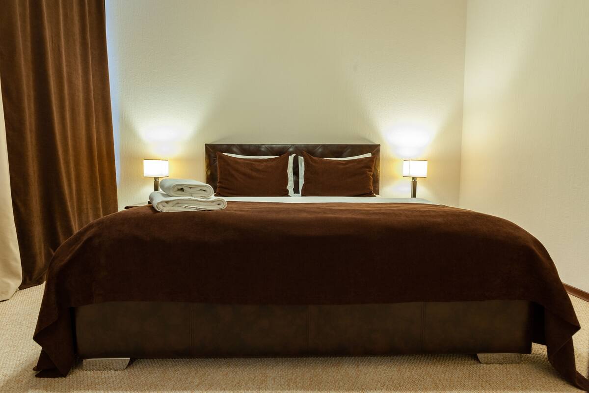 A neatly made bed with a brown bedspread, two pillows, and a folded towel on the headboard. There are two bedside lamps on either side of the bed.