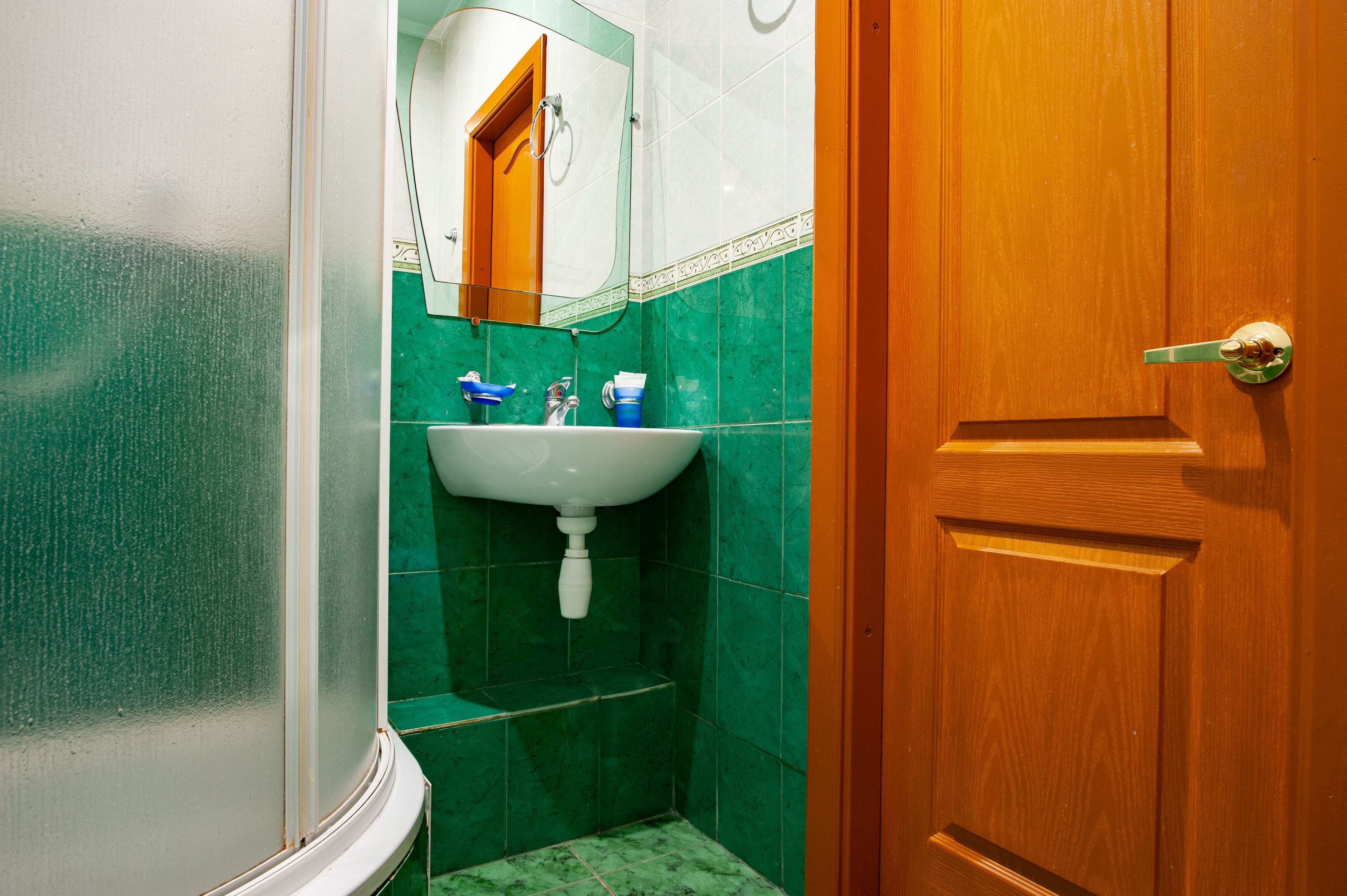 A bathroom with a white sink, green tiles, a wooden door, and a mirror above the sink.