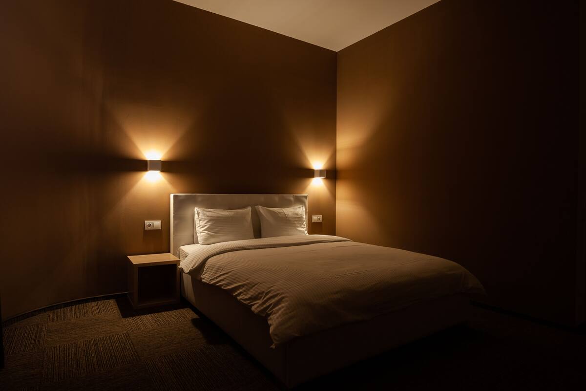 A neatly made bed in a dimly lit room with two wall-mounted lights on either side.