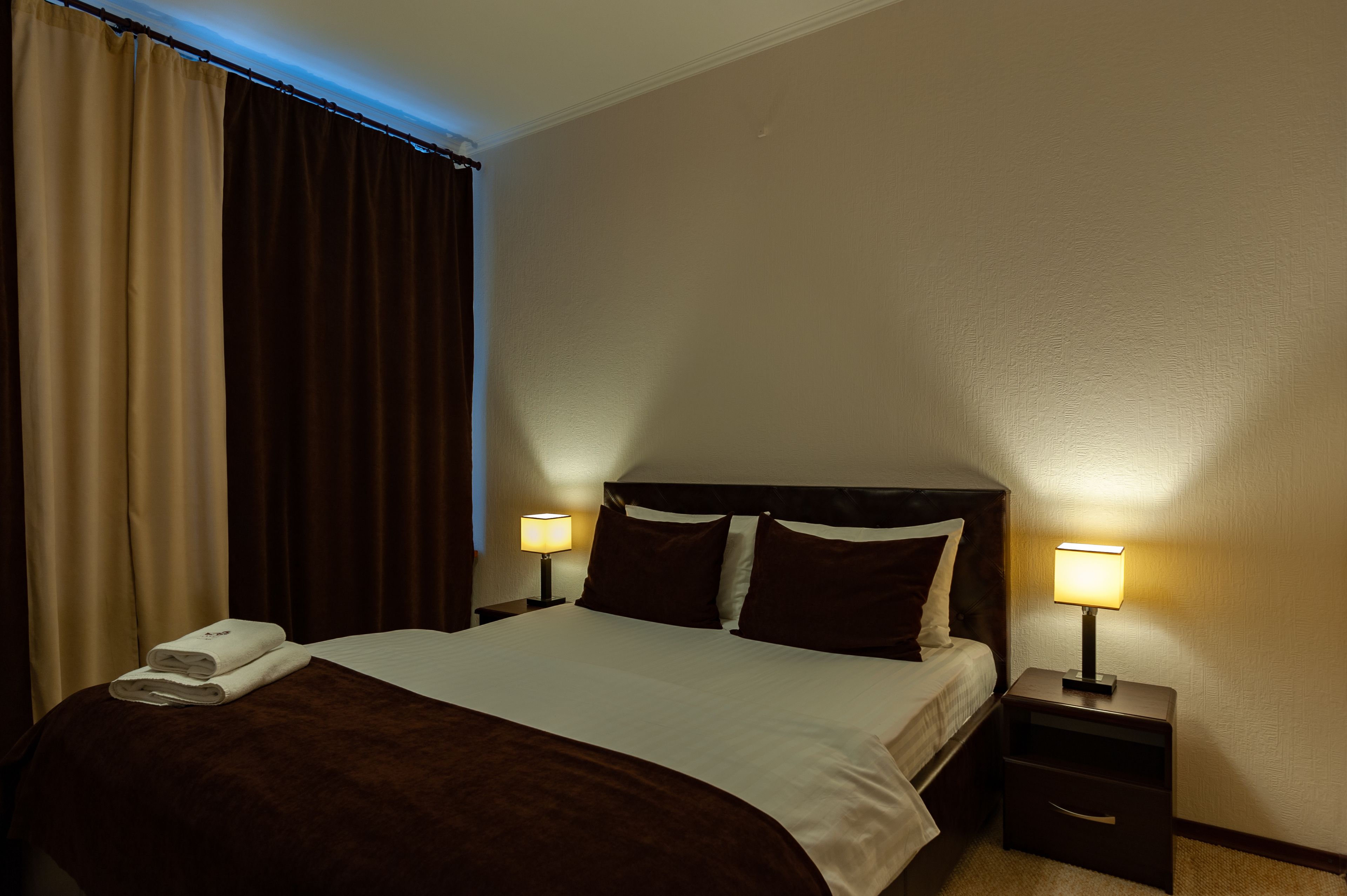 A hotel room with a bed, two bedside tables with lamps, a window with curtains, and a folded towel on the bed.