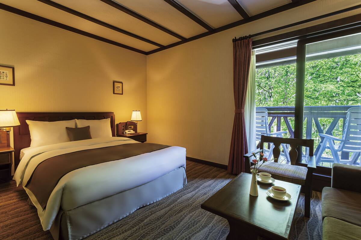Een hotelkamer met een groot bed, een bureau met stoel, een raam met uitzicht op groen en een nachtkastje met lamp.