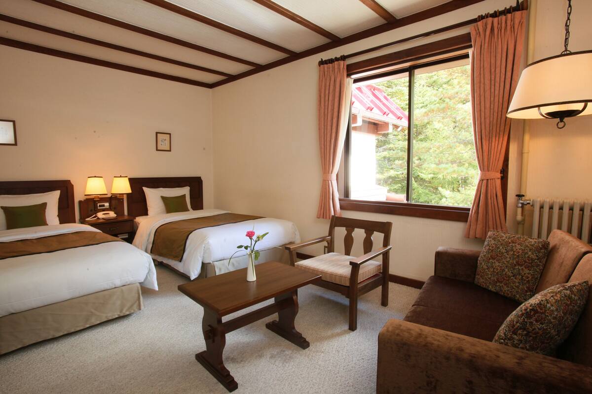 Een hotelkamer met twee bedden, een houten tafel met een bloemenvaas, een bank en een raam met gordijnen.