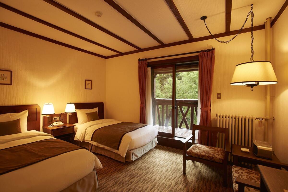 Een hotelkamer met twee bedden, een houten plafond, een raam met uitzicht, een stoel en een tafel met een lamp.