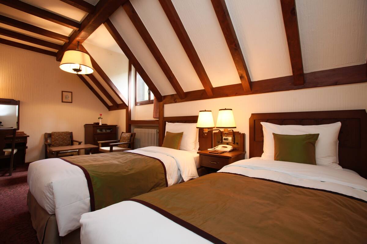 Een hotelkamer met twee bedden, houten balken in het plafond, een bureau met een stoel en een lamp.