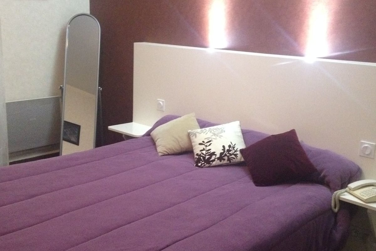 Een bed met een paars dekbed, twee kussens, een nachtkastje met een lamp en een spiegel aan de muur.