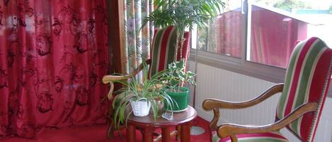 Una habitación con alfombra roja, una silla de madera con cojín a rayas, una mesita con planta en maceta y una ventana con cortinas.