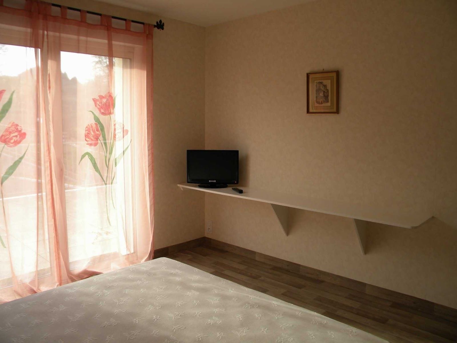 Een slaapkamer met een bed, een televisie op een plank, een raam met bloemen gordijnen, en een ingelijste foto aan de muur.