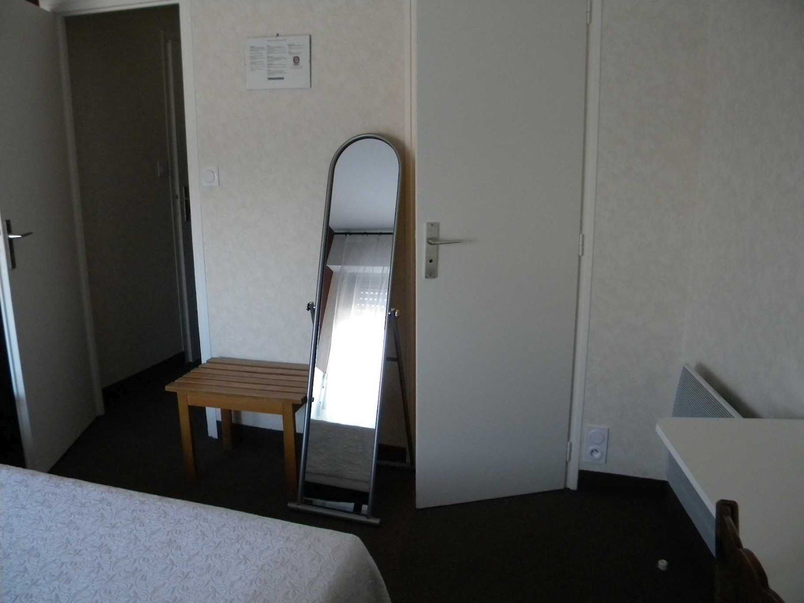 A hotel room with a bed, a small wooden bench, a mirror leaning against a door, and a white wall with a notice.