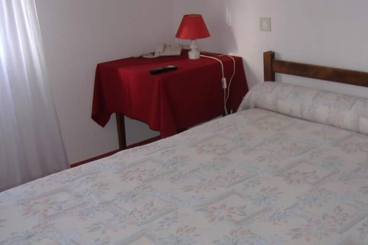 Een bed met een bloemenprint beddeken, een houten hoofdbord en een klein nachtkastje met een rode tafelkleed, een lamp en een afstandsbediening.