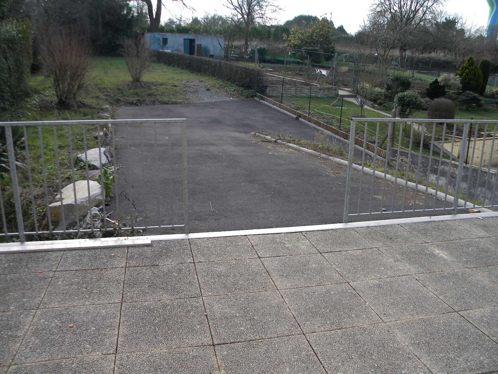 A paved courtyard with a metal fence, a stone wall, and a small building in the background.