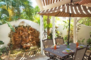 Een terras met een tafel, stoelen, een waterpartij en een pergola met een parasol.