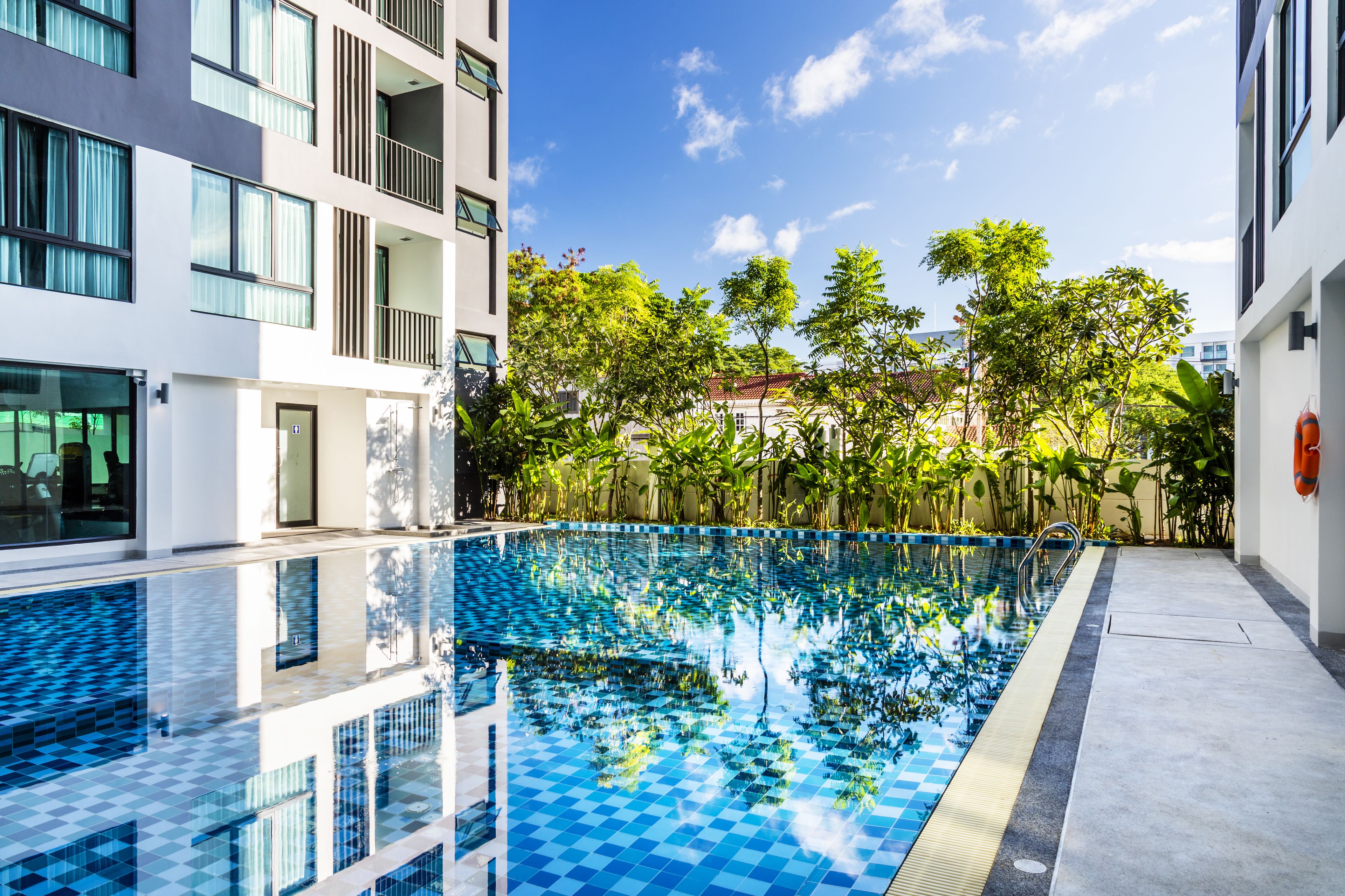 A modern building with a swimming pool, surrounded by lush greenery and a clear blue sky.