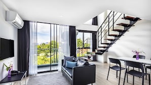 Una sala de estar moderna con un sofá de cuero negro, una mesa de comedor con sillas, una escalera y un gran ventanal con cortinas.