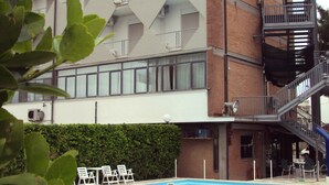 A swimming pool with lounge chairs, a brick building with a unique geometric facade, and a fire escape. - Hotel Quadrifoglio (Comacchio)