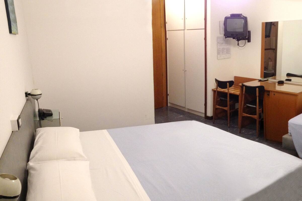 A hotel room with two single beds, a desk with a chair, a small table, a mirror, and a TV.