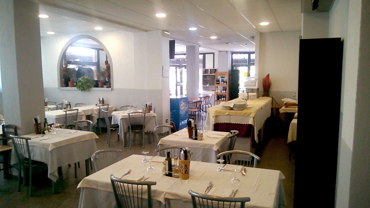 Ein Restaurant mit mehreren Tischen und Stühlen, einige davon mit weißen Tischdecken. Auf den Tischen stehen Flaschen und Gläser, im Fenster steht eine Pflanze.