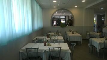 Zona pranzo con diversi tavoli e sedie, un'ampia finestra con tende e uno specchio sopra i tavoli.