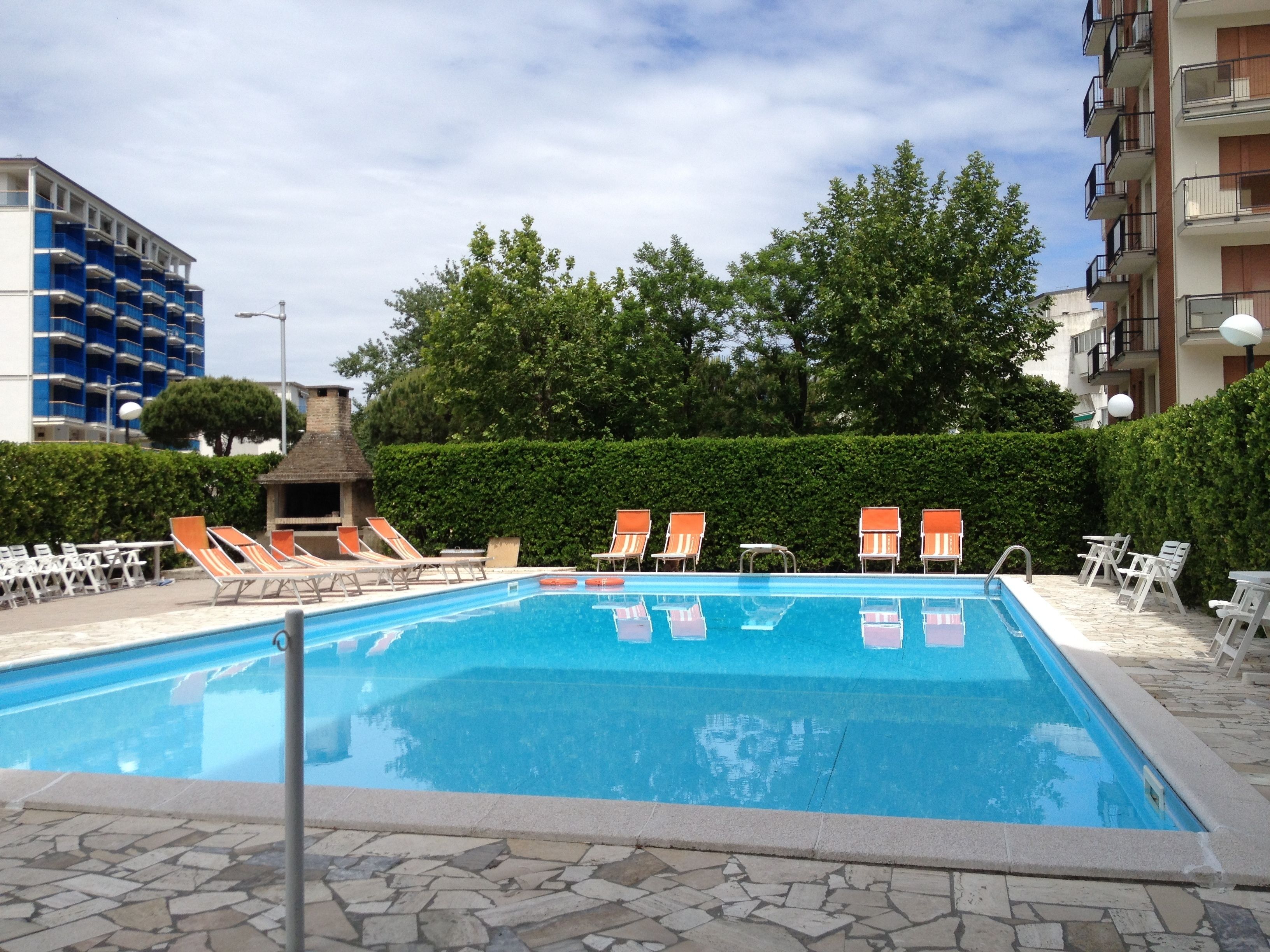 A swimming pool with lounge chairs and a small fire pit area, surrounded by a hedge and trees.