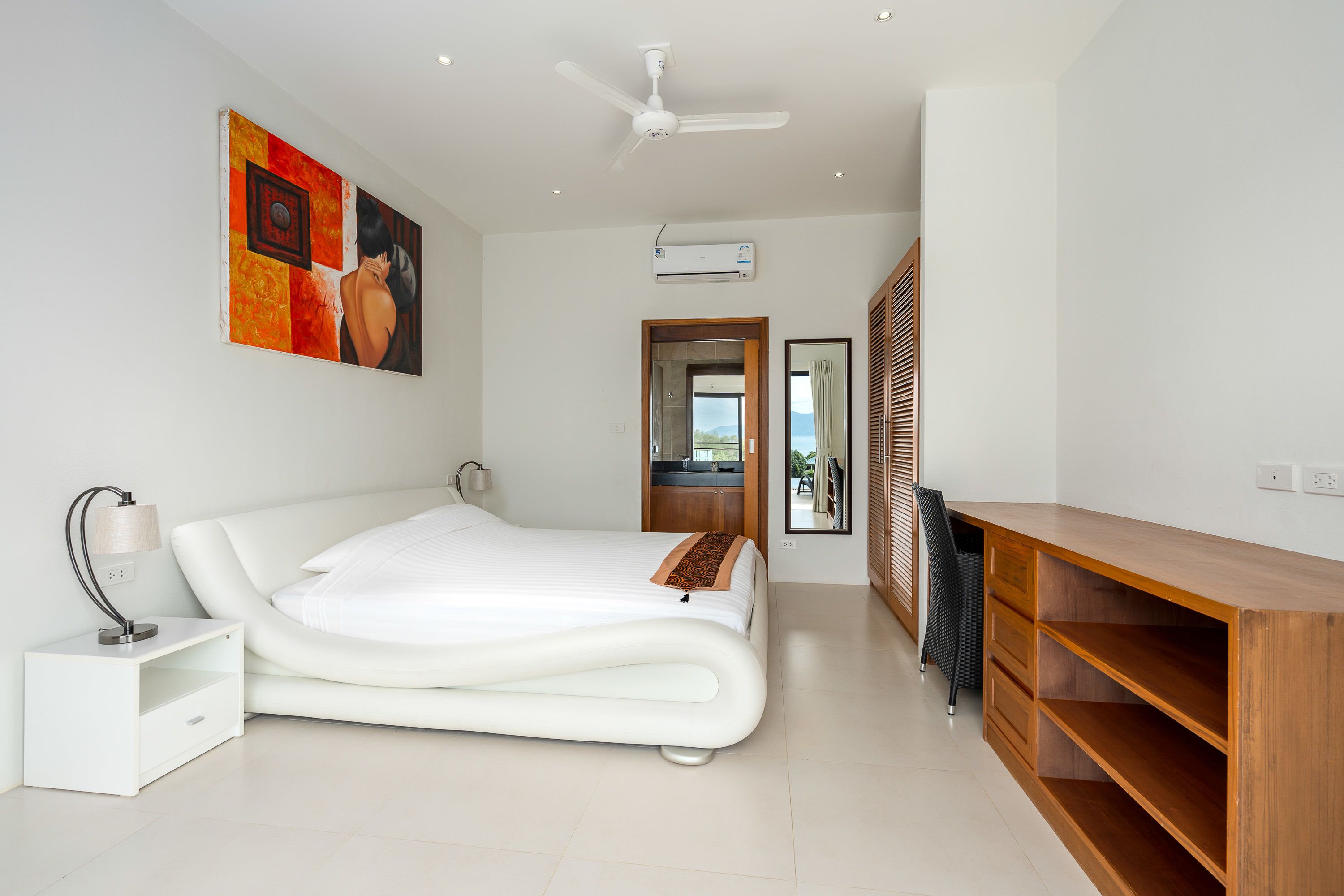 A modern bedroom with a large bed, a bedside table, a desk, and a ceiling fan.