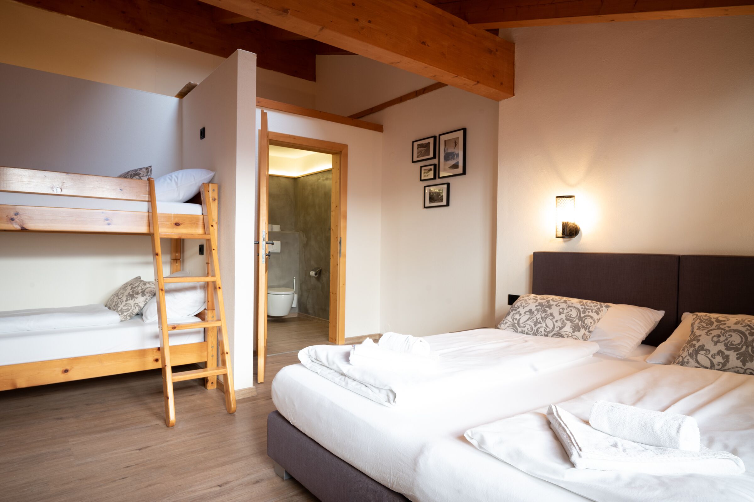 A bedroom with a bunk bed, a separate bed, a wooden ladder, a bathroom door, and a wall-mounted light fixture.