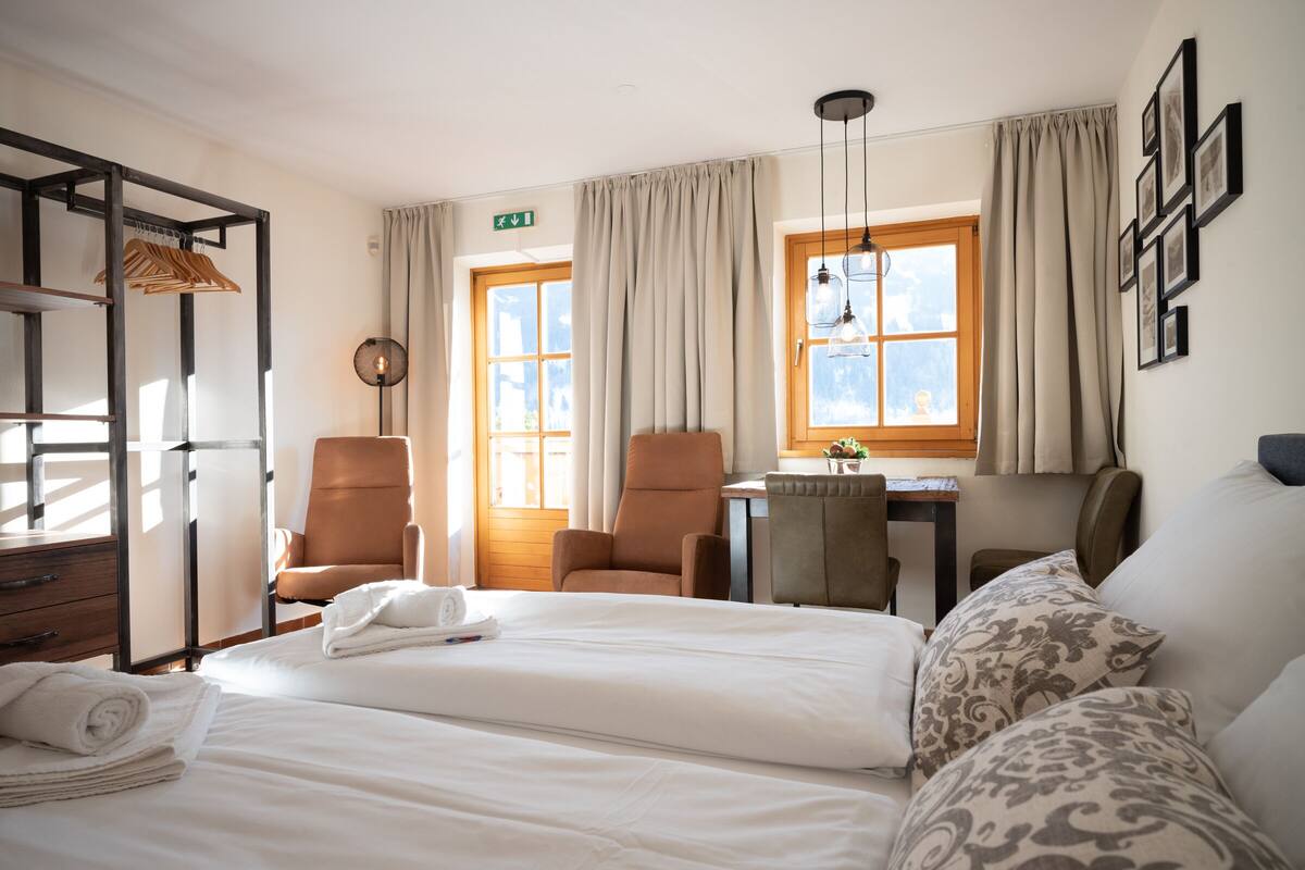 Une chambre d’hôtel avec un lit, deux chaises, un bureau, un luminaire suspendu et une fenêtre avec des rideaux.