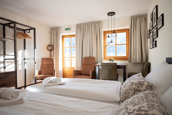 Habitación de hotel con cama, dos sillas, escritorio, lámpara colgante y ventana con cortinas.