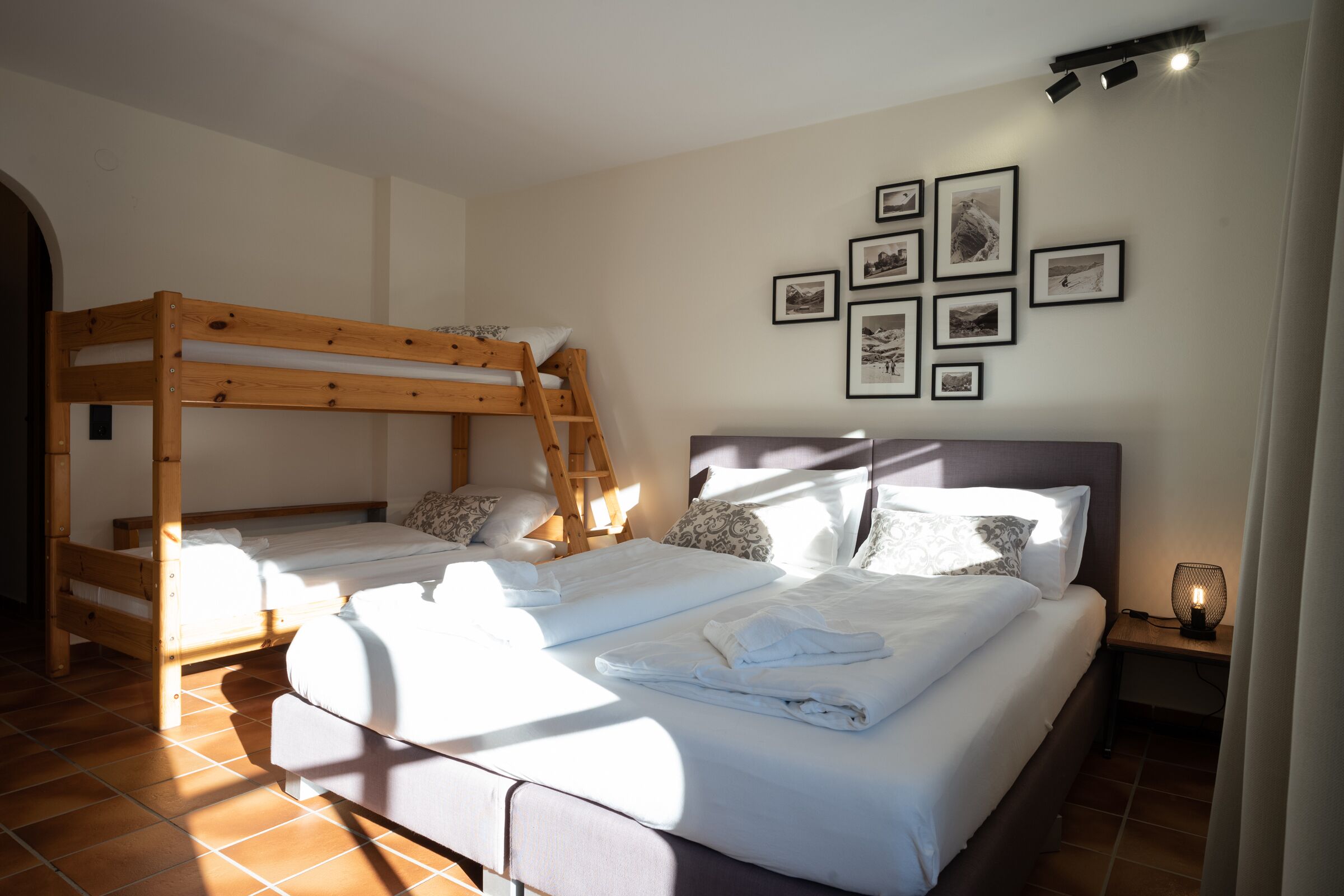 A room with a bunk bed, a single bed, and a bedside table with a lamp. There are framed pictures on the wall.