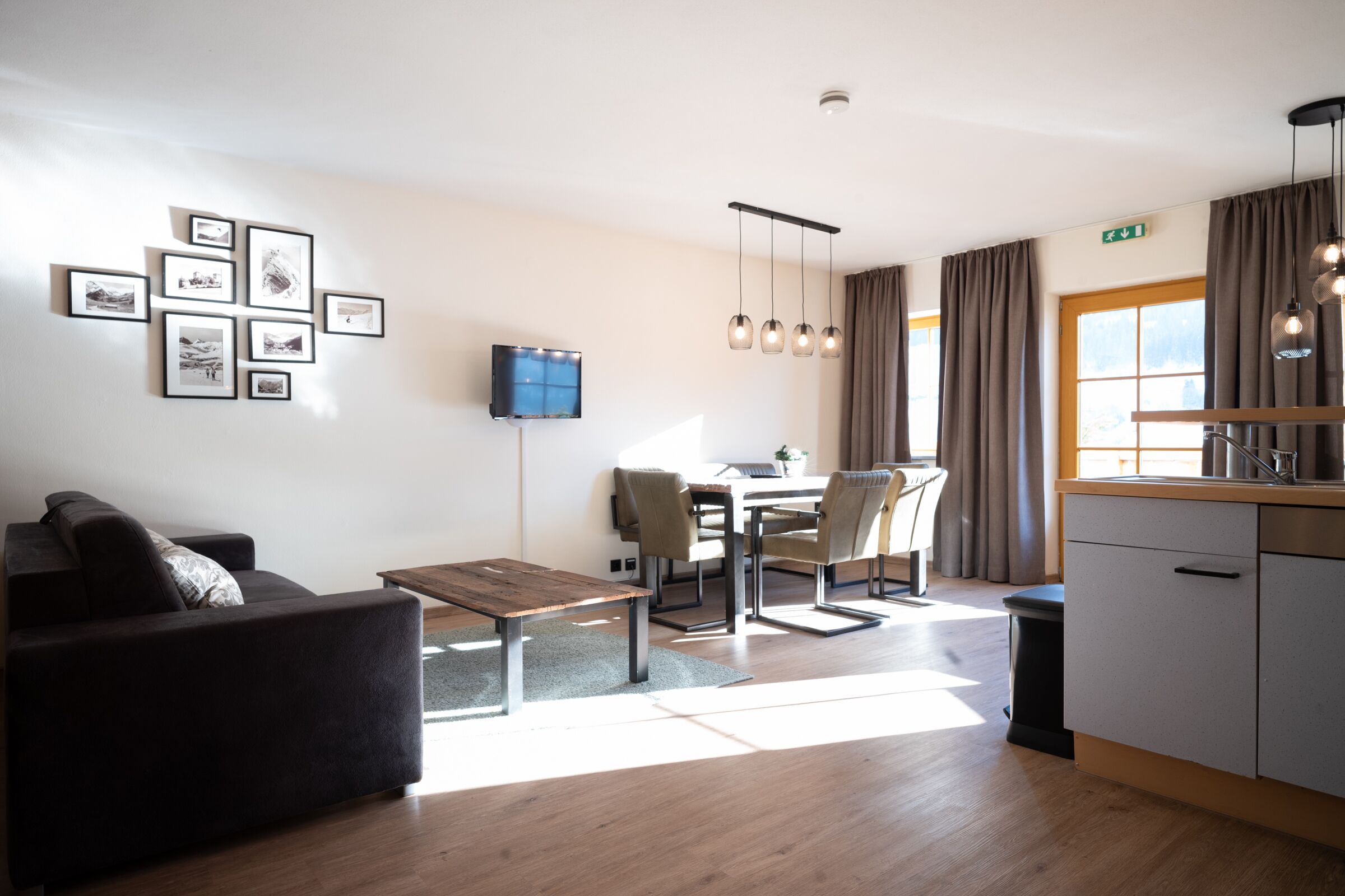 A modern hotel room with a sofa, dining table, chairs, a TV, pendant lights, a kitchen area, and a large window with curtains.