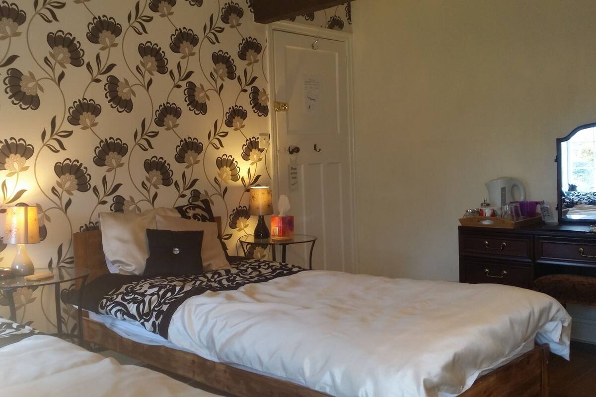 Quarto com duas camas, papel de parede floral, luminárias de cabeceira, uma cabeceira de madeira, uma mesa pequena com abajur e um penteado com gavetas.