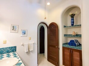 Un baño con lavamanos blanco, salpicadero de azulejos azul y blanco, puerta de madera y repisa con toallas y un jarrón.