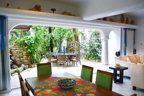 Un espacio de sala de estar integrado con una mesa de comedor, sillas y un sofá. Se ve un jardín exuberante a través de un arco.