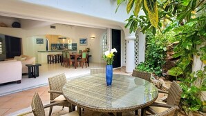 A spacious living area with a round glass table and wicker chairs, a dining area with a white table and chairs, and a kitchen visible in the background. - Natz Ti Ha H101 by PRP (Playa del Carmen)