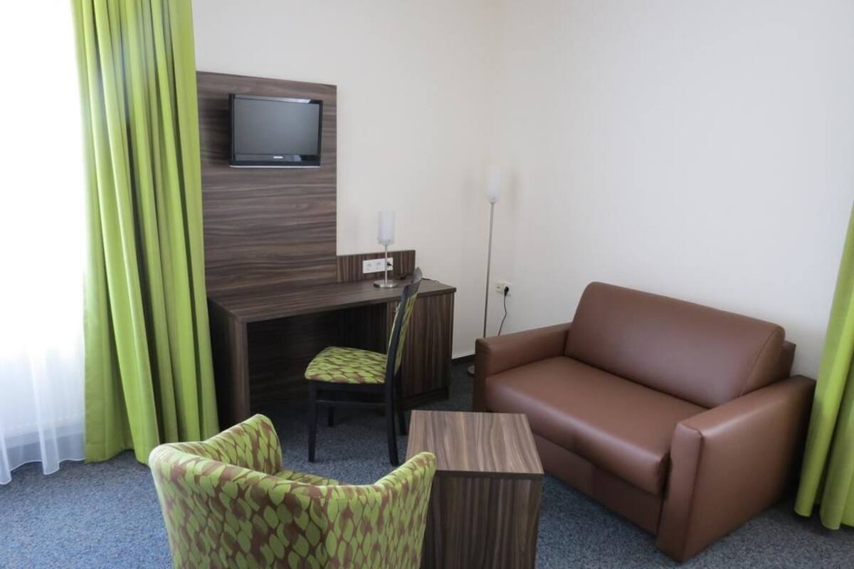 A hotel room with a brown leather sofa, a green patterned armchair, a wooden coffee table, a desk with a chair, and a flat-screen TV mounted on the wall.