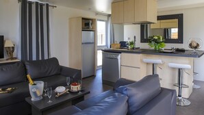 A modern living room with a sofa, a dining table with a bottle of champagne, and a kitchen area with a refrigerator, dishwasher, and bar stools. - Camping Le Clos du Rhône (Saintes-Maries-de-la-Mer)
