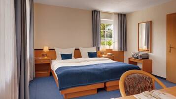 A hotel room with a bed, bedside tables, a chair, a desk, a lamp, a suitcase, and a window with curtains.