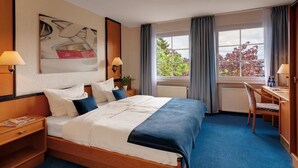 Habitación de hotel con una cama grande, dos mesitas de noche con lámparas, un escritorio con silla y una ventana con cortinas.